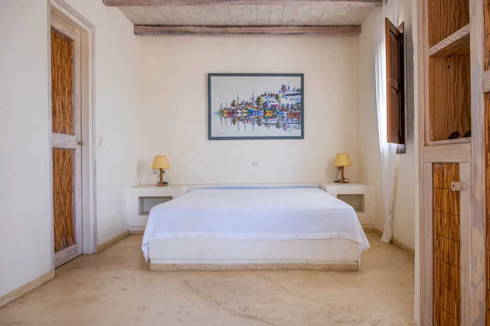 Double bed on a low plaster platform, whitewashed walls, a harbour painting above, and two yellow bedside lamps