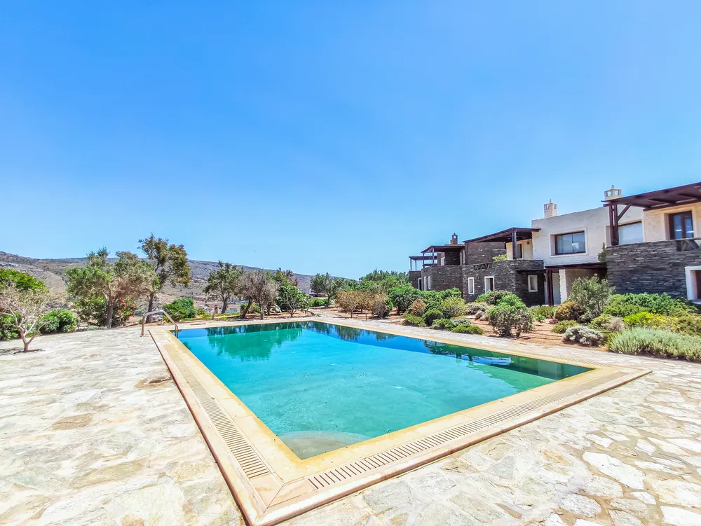 Pool terrace in bright midday sun, with the two-storey stone villa and Mediterranean shrubs behind